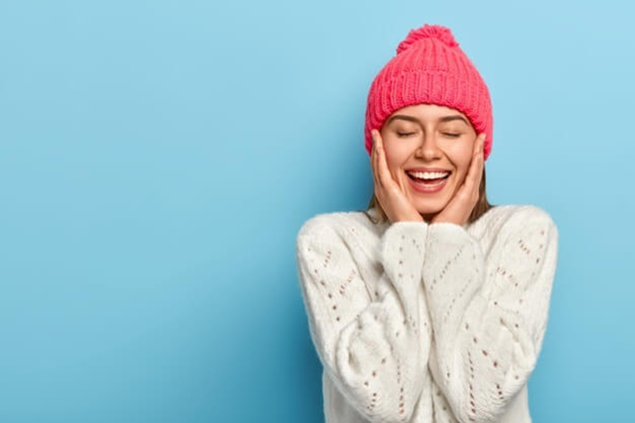 charming smiling feminine girl touches cheeks with both hands eyes shut toothy alluring smile wears winter white sweater poses against blue studio wall has pure healthy skin feels relieved 273609 32 - مقالات